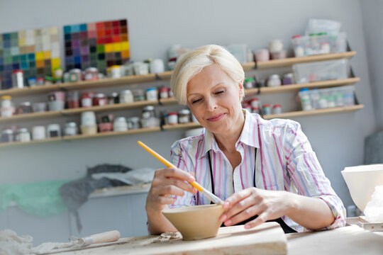 Smiling mature woman painting pottery bowl in studio