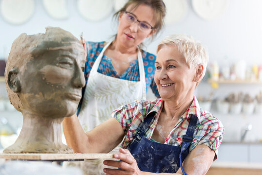 Teacher guiding senior woman sculpting clay face in pottery studio
