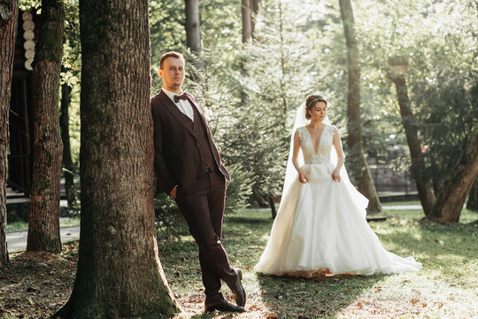 full-length portrait of a young bride and groom walking on a green golf course at sunset. Happy wedding couple, space for text. The groom leans against a tree, the bride is behind - Powered by Adobe
