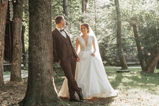 full-length portrait of a young bride and groom walking on a green golf course at sunset. Happy wedding couple, space for text. The groom leans against a tree, the bride is behind - Powered by Adobe