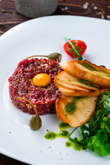 Beef tartare with raw egg, fried bread, capers, French mustard, tomatoes, fresh salad and sauce in a plate.