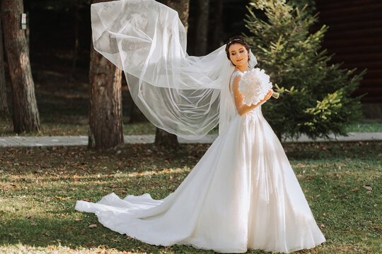 Full length side view of one beautiful sensual young brunette bride in long white wedding dress and veil standing in park holding bouquet outdoors on natural background, horizontal picture