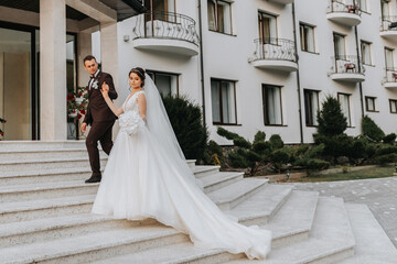 Fototapeta premium Groom and bride walking outdoors near a posh hotel. A long train of dress on the steps