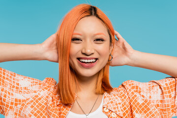 positivity, radiant smile, young asian woman with dyed hair standing in orange shirt and smiling on blue background, casual attire, freedom, cheerful attitude, looking at camera
