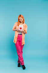 full length of young and joyful asian woman with happy smile, in trendy casual clothes standing with gift box on blue background, dyed red hair, orange shirt, pink pants, modern fashion