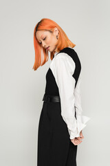 side view of expressive and young asian woman posing with hands behind back on grey background, dyed red hair, white shirt, vest and pants, business casual fashion, modern lifestyle