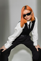 modern fashion shoot, trendy and red haired asian woman in dark sunglasses and black and white business casual clothes standing in stylish pose on grey shaded background, generation z