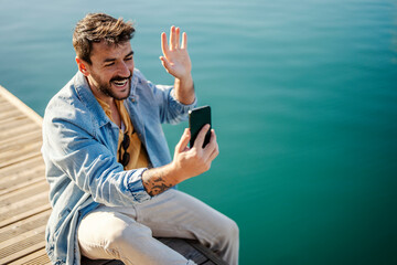 A friendly man is sitting on the dock and having video call on the phone.
