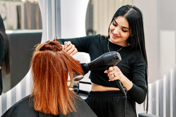Beautiful woman stylist doing hair styling with hairdryer at client in modern beauty salon....