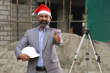 Hardworking confident Indian Site engineer figuring out the work in progress on Xmas Day. On the site location wearing the uniform and holding helmet in white. Wearing a Santa hat he is at work 