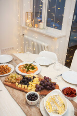Festive celebration table decorated with white and golden tablecloth, with salad assortment, cheese platter, smoked salmon, wine glasses, bokeh lights