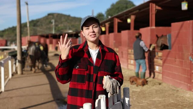 Portrait of positive talking European young woman wearing plaid shirt and posing on yard of horse stables