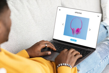 African american man sitting on sofa with modern laptop with stylish headphones on screen, buying at online store
