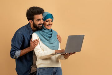 Young muslim couple using laptop computer while standing over beige studio background
