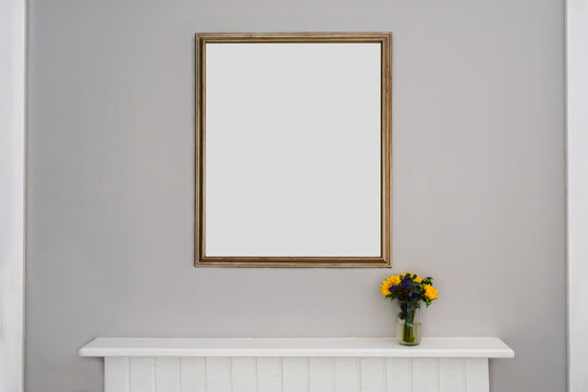 A gold frame hangs on a simple, moden grey wall with a vase of flowers sitting on the top of a fireplace