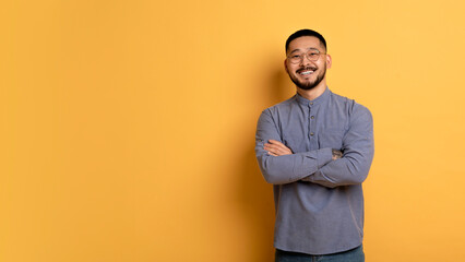 Confident young asian man standing with folded arms over yellow background
