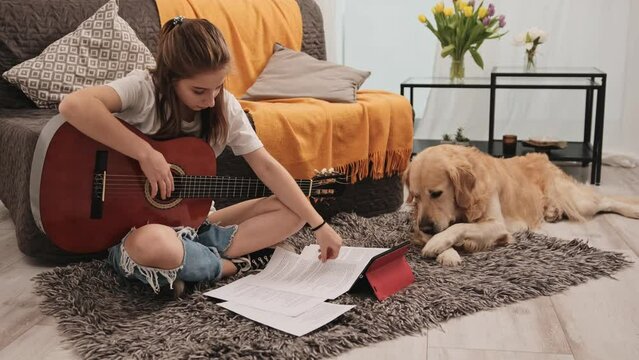 Beautiful young girl teenager practicing guitar at home and golden retriever dog pet lying on floor. Musical instrument playing lesson at living room with purebred doggy