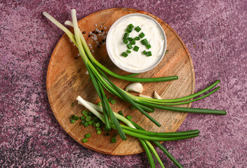 Board with bowl of tasty sour cream and sliced green onion on purple background