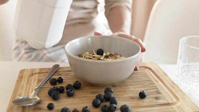 Girl puts milk to oatmeal with blueberry in bowl plate for delicious cereal breakfast at kitchen. Woman preparing granola with berry and latte for morning healthy diet meal