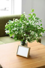 Vase with blooming jasmine flowers  and blank frame on wooden coffee table in living room
