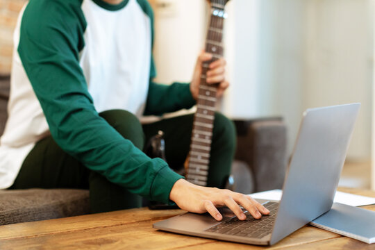 Cropped Shot Of Guy With Guitar Typing On Laptop Indoors