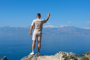 Fototapeta na wymiar Man stands waving hand greeting on rock blue ocean mountain peaks horizon. Active holiday adventure, tourism action, healthy summer joy, Fun activity lifestyle. Crazy adult guy fly from climb.