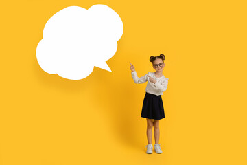 Smiling Little Schoolgirl Pointing Aside At Empty Speech Bubble Over Yellow Background