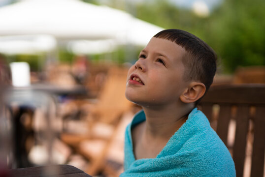 A 7-year-old blond boy sits on a lounger with a towel on his shoulders. The child recently got out of the pool.