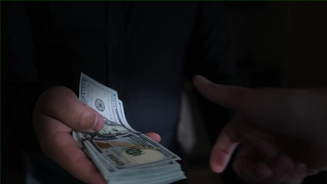 Two men are exchanging money, the man's hand abruptly gives a large wad of cash dollars to the other person in the palm. Close up on dark background, vignette. Bribery and extortion of money concept. 
