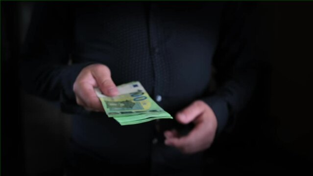 Man's hand holds out stack of one hundred euros bills to another person, close-up on blurred dark background. Significance of trust and professionalism in business dealings.