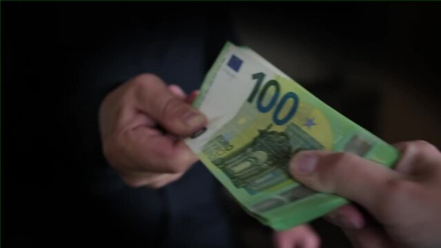 Two people are trying to divide money among themselves in dark room, man's hand is pulling pack of 100 euro bills strongly, close up, black vignette.
