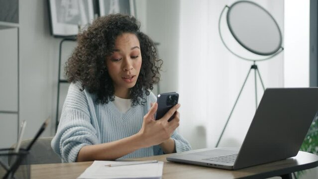 Remote work at home office. Productive businesswoman sitting at desk working on laptop and talking by video call looking at smartphone camera. Mixed race positive free lance woman entrepreneur at work