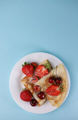 delicious fragrant pancakes folded in a triangle on a white plate and a blue background and decorated with delicious ripe sweet strawberries, red fragrant cherries and coconut flakes
