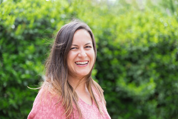 Fototapeta premium Outdoor portrait of a laughing 45 year old woman with windblown brown hair looking at the camera in the park on a sunny day. Confidence and tranquility of a mature woman. Happiness, lifestyle.