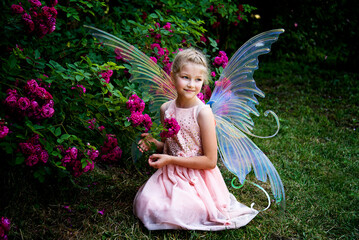 little beautiful girl in a fairy costume of butterfly with wings sitting on the grass in roses garden on sunny summer day 