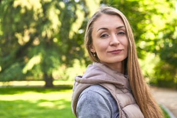Obraz premium Radiant middle-aged Caucasian woman, smiling in the morning sunlight at the park, embracing the beauty of nature and finding joy in peaceful serenity