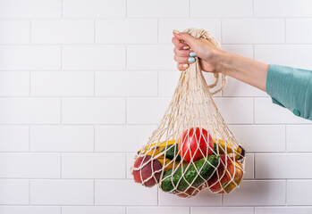 Woman's hand holding mesh string bag full of fruits and vegetables. International plastic free day background, shopping concept. 3rd of July. Copy space