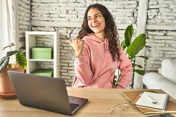 Young student woman studying in her home points with thumb finger away, laughing and carefree.
