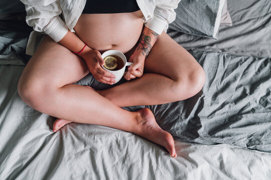 Young pregnant woman sitting with cross-legged on the bed. Cup of tea close up photo in bedroom in the early morning time. Woman's health, happy pregnancy and calm mental mood concept image.