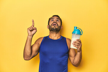 Athletic young Latino man holding a protein shake against a yellow background pointing upside with...