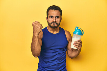 Athletic young Latino man holding a protein shake against a yellow background showing fist to camera, aggressive facial expression.