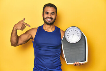 Fit young Latino man with scale on yellow background, fitness concept, person pointing by hand to a shirt copy space, proud and confident