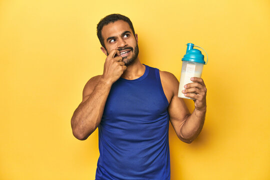 Athletic young Latino man holding a protein shake against a yellow background relaxed thinking about something looking at a copy space.