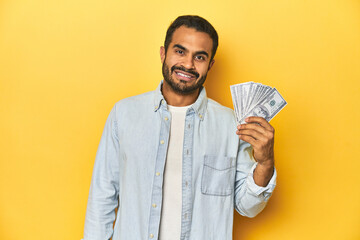 Young Latino man holding a bundle of dollars, yellow studio background, happy, smiling and cheerful.