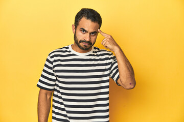 Casual young Latino man against a vibrant yellow studio background, pointing temple with finger, thinking, focused on a task.
