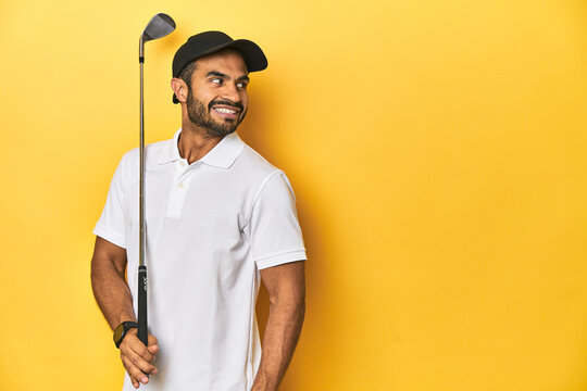 Young Latino golfer with club and cap on a yellow studio background, looks aside smiling, cheerful and pleasant.