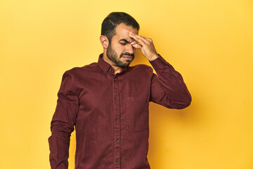 Casual young Latino man against a vibrant yellow studio background, having a head ache, touching front of the face.