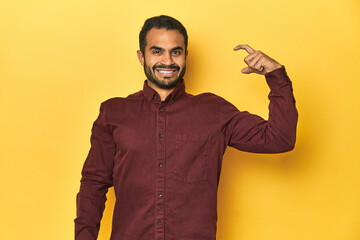 Casual young Latino man against a vibrant yellow studio background, holding something little with forefingers, smiling and confident.