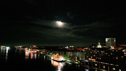 Drone photo at night over the Savannah River. Flying over ferry boat a brightly lit boardwalk with moon in distance.