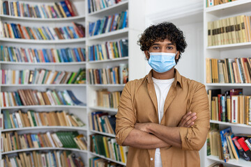 Portrait of young student inside library, man with crossed arms looking at camera, hispanic man with protective face mask visiting academic library and bookstore.